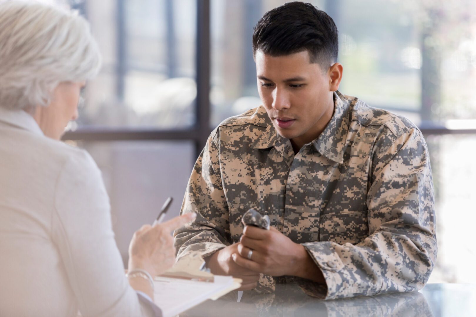 Mid-adult male in military uniform anxiously clenches his camouflage hat in his hands as he sits down at a table with female counselor. She holds a clipboard and pen as she gestures at him with her finger.